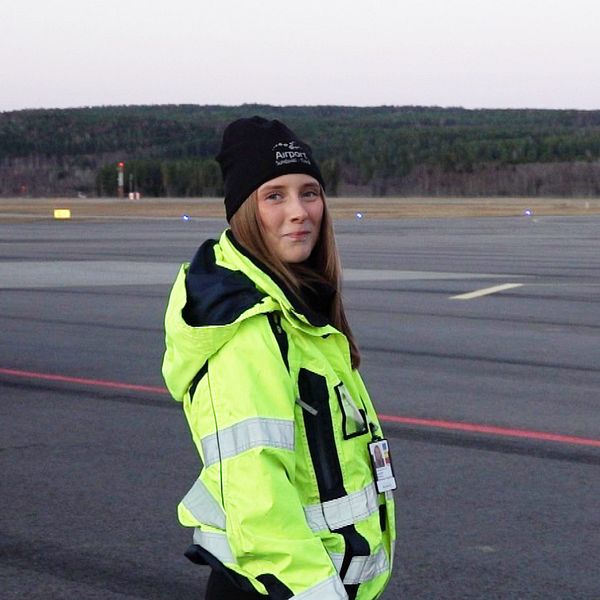 En person i en gul och svart västs övervakning av landningen av ett flygplan på en flygplats under skymning. Flygplanet ankommer på flygplansbanan i bakgrunden, omgiven av träd.