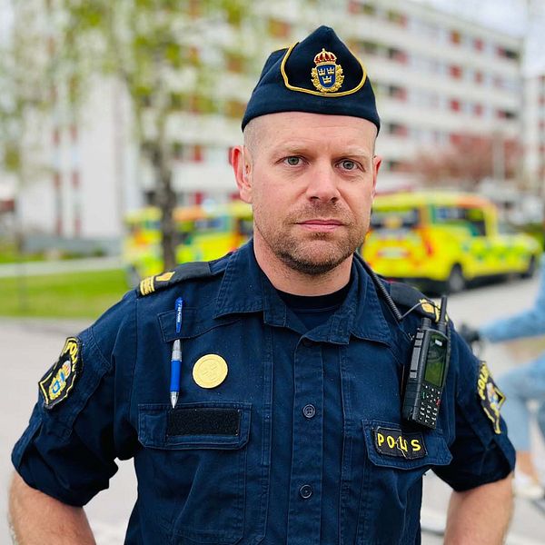 En polis står i ett bostadsområde, i bakgrunden syns blåljusfordon. Till höger syns brandbilens stegbil och en ambulans.