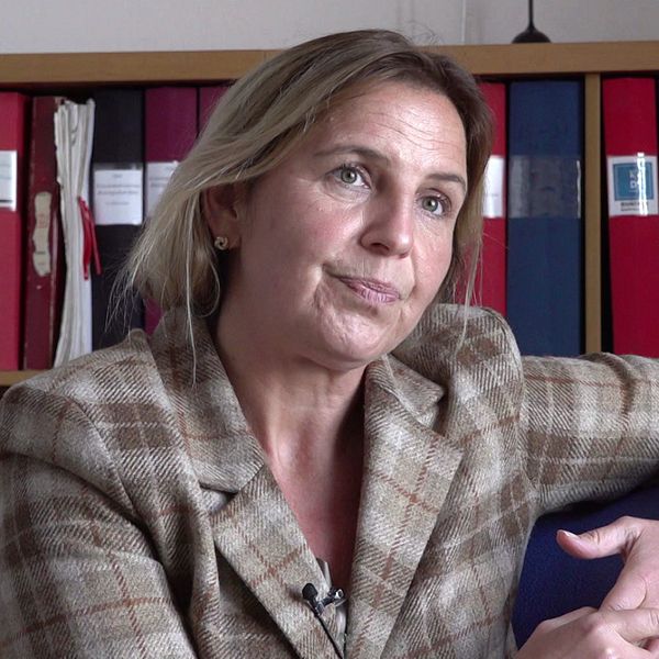 En delad bild som visar två personer som deltar i en intervju. Den vänstra sektionen visar en kvinna i en rutmönstrad kavaj som sitter i en stol, omgiven av en biblioteksliknande miljö med arkivlådor i bakgrunden. Den högra sektionen visar en man klädd i en svart väst som sitter med armarna i kors, framför en neutral bakgrund.