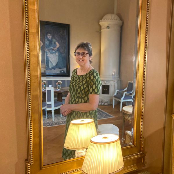 Landshövding Brittis Benzler står i sitt hem och tittar in i kameran via en spegel med förgylld ram.