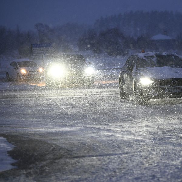 En bil kör i snöigt och dimmigt väder, med snöflingor som virvlar runt. Två andra bilar syns i bakgrunden med sina strålkastare tända. Känsla av en kall vinterkväll.