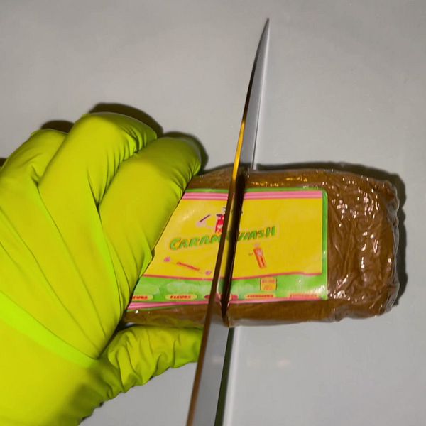 En handskklädd hand och en kniv som skär i ett paket med narkotika och en annan handskklädd hand som håller i en påse med vitt pulver.