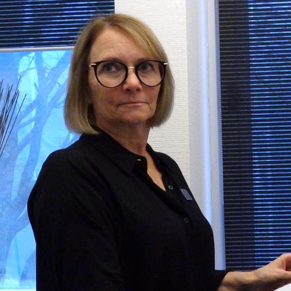 En kvinna står vid en dator i ett rum med fönster som har persienner. Utomhus syns trädgrenar och en blåaktig himmel. Personen bär en svart skjorta.