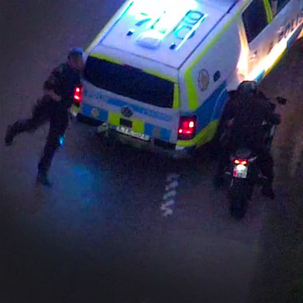 Polisbil som har stängt vägen för en motorcyklist och en polisman springer rund bilen för att gripa en man som sitter på en motorcykel. Sett från polisen helikopter.