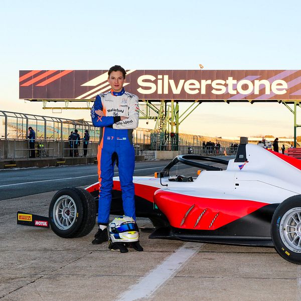 En ung förare står bredvid en racingbil på Silverstone-banan, med en stor skylt som visar banans namn i bakgrunden. Föraren är klädd i en blå och orange racingdräkt och håller en hjälm i handen. Racingbilen är vit med röda och svarta detaljer och har nummer 9 på sidan.