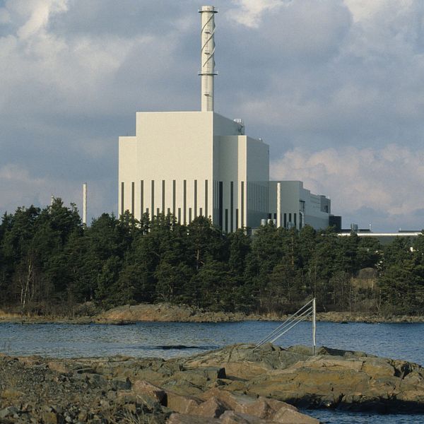 Kärnkraftverk vid kusten, omgiven av vatten och skog under molnig himmel.