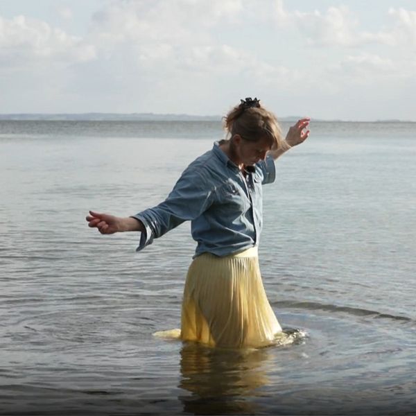 En delad bild med två sektioner. Den vänstra sektionen visar en person som går i grunt vatten vid havet, klädd i en blå skjorta och en gul kjol, med armen utsträckt. Den högra sektionen visar samma person stående inomhus vid ett bord, omgiven av bilder av olika fiskslag på väggen.
