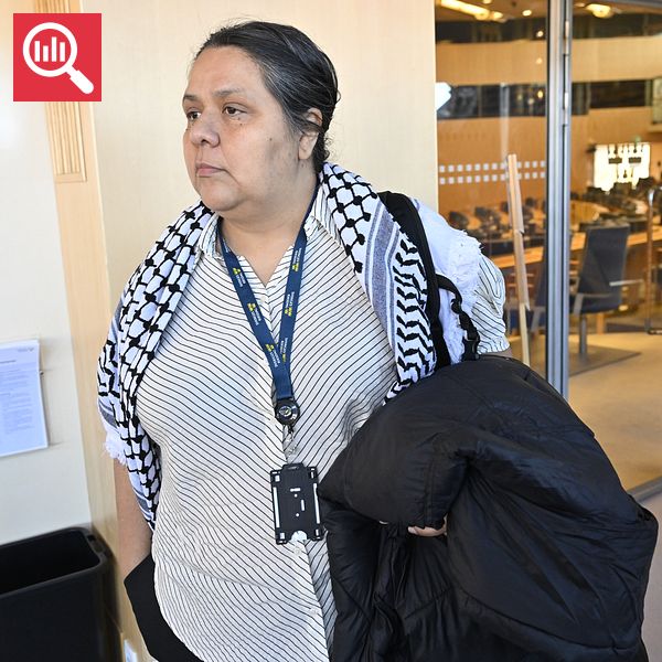 Person med palestinasjal står med jacka över armen, riksdagens plenisal och fler personer syns i bakgrunden.