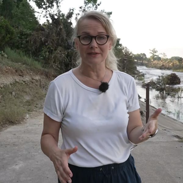 Reporter vid översvämmad flod, bild på bråte och förstörda fordon i vattnet.