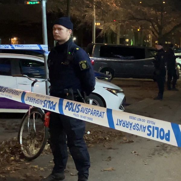 Poliser står vid en avspärrning på en gata, omgiven av flera fordon. Bredvid en cykel är avspärrningsbandet tydligt synligt med texten ”POLIS AVSPÄRRAT”. I bakgrunden syns fler poliser som arbetar i området, under en mörk kväll.
