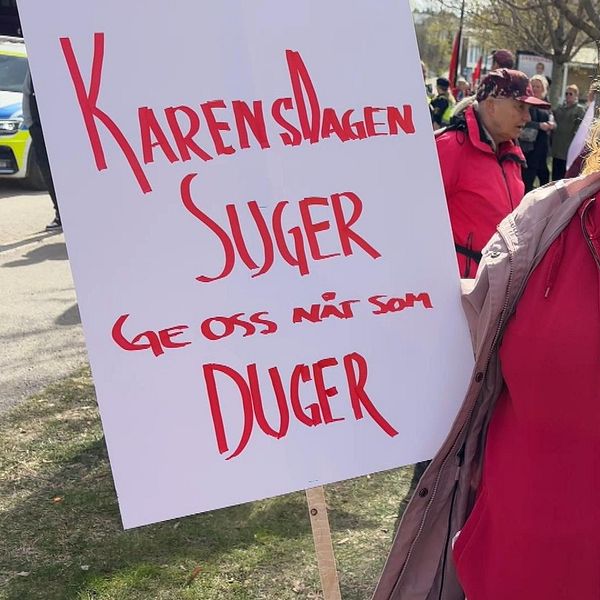 Demonstrant i röd jacka håller skylt med texten ”Karensdagen suger. Ge oss nåt som duger”.