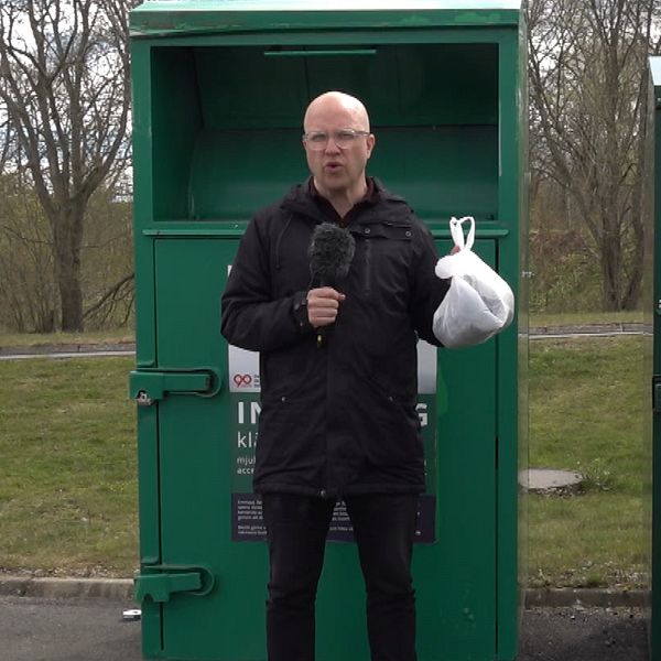 En reporter håller upp en plastpåse med kläder i. Han står framför gröna insamlingsboxar.