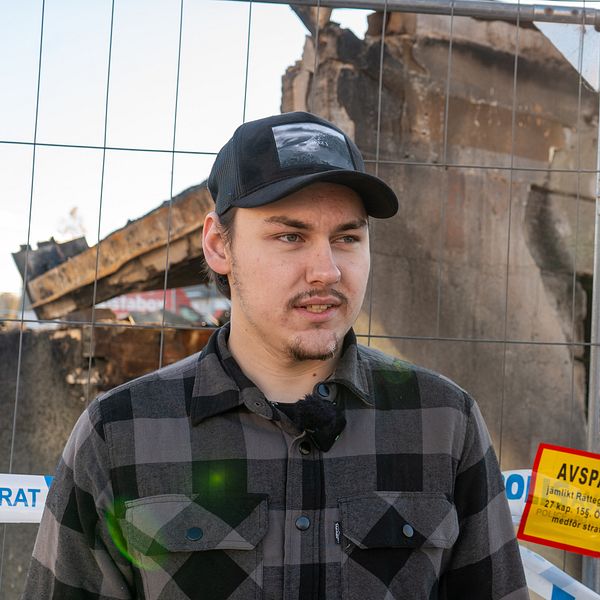 Alvar Claesson står framför en avspärrning vid en brandskadad byggnad. I bakgrunden syns resterna av byggnaden med tydliga skador, och en solstråle bryter genom den krisande strukturen. En röd och gul avspärrningsskylt är placerad i bildens nedre del.