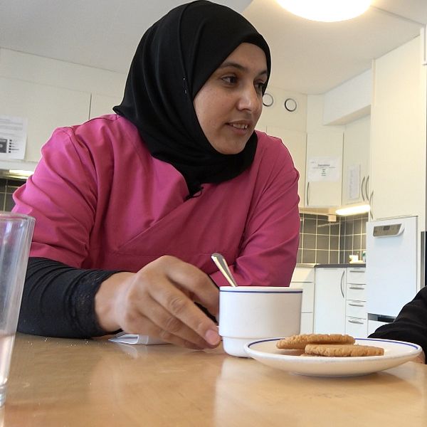 En kvinna i sjal och rosa tröja serverar en äldre kvinna kaffe och kakor vid ett bord