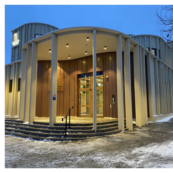 Till vänster syns Norrköpings tingsrätt. Byggnaden är omgiven av snö och grus. Till höger står en man som är klädd i en mörk tröja, positionerad på en inomhustrappa i en ljus miljö.