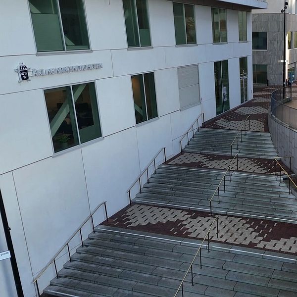 Trappor som leder ner från en byggnad med arkitektonisk design, omgivna av en innegård och andra byggnader. En bil är parkerad i bakgrunden.