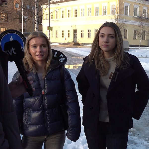 Tre tjejer står på en snötäckt gata, klädda i vinterjackor, med en byggnad i bakgrunden och en färgglad muralmålning.