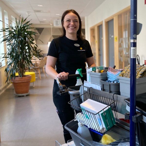 En lokalvårdare skjuter en städvagn med rengöringsprodukter och redskap i en korridor. Till vänster syns en stor krukväxt och till höger en dörr med skylten ”Endast personal”.