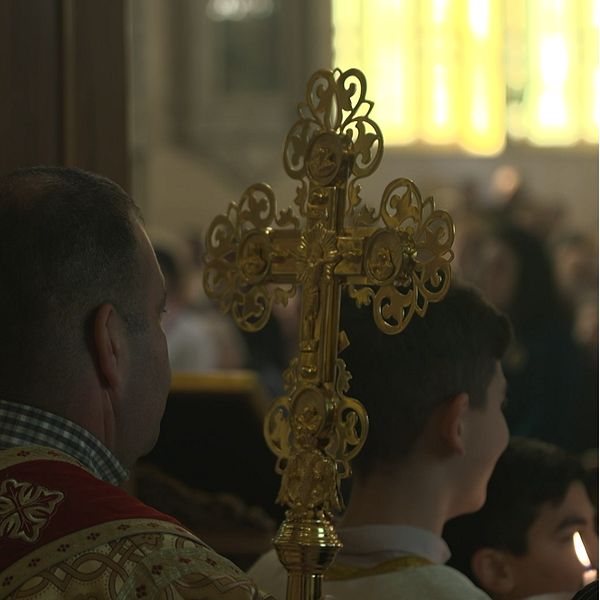 Bilden är delad i två sektioner. Den vänstra sektionen visar en guldutsmyckad kors som hålls av en man under en religiös ceremoni, med en liten pojke synlig i bakgrunden. Den högra sektionen visar en kvinna med långt, lockigt hår som står utomhus med en klar himmel i bakgrunden.