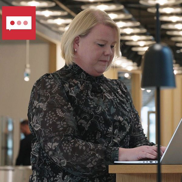 En person står vid en stående arbetsstation och arbetar på en laptop i en modern, ljus arbetsmiljö. I bakgrunden syns en kollega sittande vid ett bord. Rummet är väl upplyst med moderna lampor och har trämaterialinslag.
