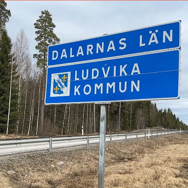 En  typisk svensk landsbygdsscen med en vägskylt som markerar övergången till Dalarnas Län och Ludvika Kommun. Skylten, som är blå med vit text och det gula Dalarnas landskapsvapen, står vid en vägkant. I bakgrunden syns ett kalt trädlandskap under en grå himmel, vilket antyder att det är sent på hösten eller tidigt på våren. Till höger i bilden finns en person klädd i en blommig blus och mörk jacka, stående framför ett rött hus med vita knutar.  Det är Ljusnarsbergs kommunalråd Ewa-Leena Johansson (S).