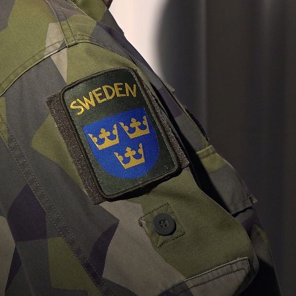 Närbild på ärmen på en person i militärkläder, skuggan av honom syns på väggen bakom.