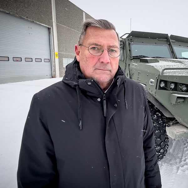 En person står i snöigt landskap framför en grön militärfordon med en robust design. Fordonet har stora däck och en kraftig framgrill, och i bakgrunden syns en byggnad och snöklädda träd.