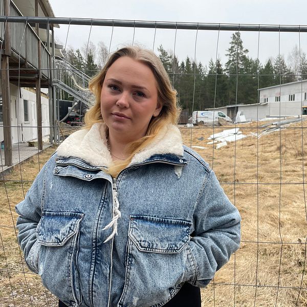 Person i jeansjacka framför ett stängsel med byggnader i bakgrunden.
