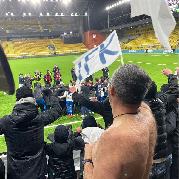 En delad bild som visar två scenarier från en fotbollsmatch. Den vänstra sektionen visar en grupp entusiastiska supportrar med en flagga som hyllar sitt lag, medan spelarna firar i bakgrunden. Den högra sektionen visar en ensam supporter som poserar utan tröja, med en gest av seger och en pins från laget. Stadion är delvis upptagen med åskådare.