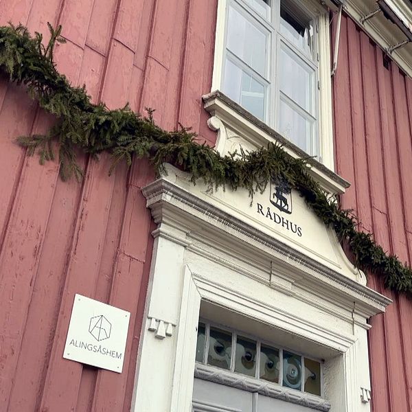 Bildens vänstra sektion visar en del av ett rött träbyggnad som är dekorerat med girlander och har en skylt med texten ”RÅDHUS” ovanför en dörr. Det finns också en annan skylt märkt ”KUNGSBACKA” närmare dörren.