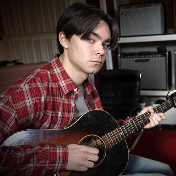Person i röd skjorta spelar gitarr omgiven av förstärkare och musikinstrument.