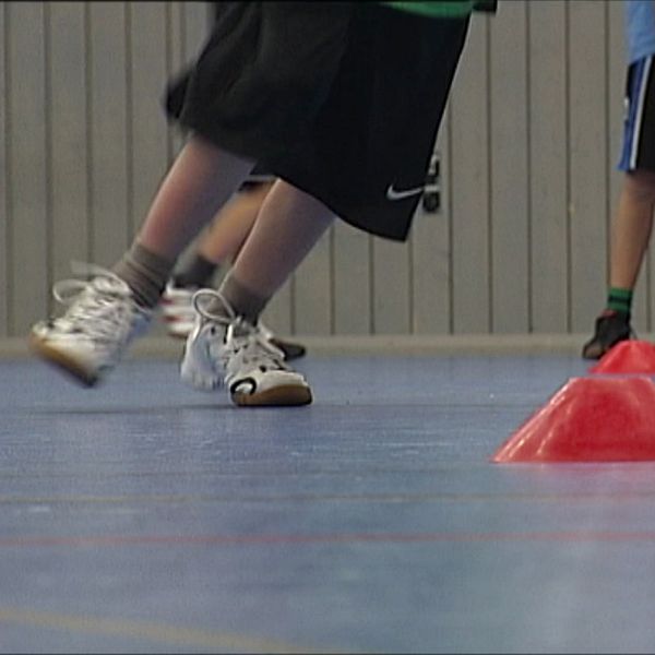 Aktiva barn som spelar inomhus, rör sig kring orange koner på ett gymnastikgolv. Fokus ligger på benen som springer mot konerna, medan andra barn syns i bakgrunden som deltar i aktiviteten. Det handlar om att fler barn och unga i Västerbotten får fysisk aktivitet på recept.