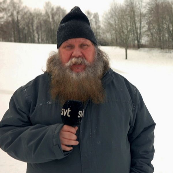 Vinterklädd reporter med stort skägg håller i mikrofon i ett kyligt vinterlandskap.