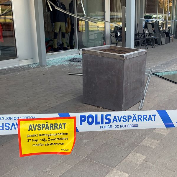 Avspärrningsband framför butik, krossat glas ligger på marken och en polisbil står parkerad i bakgrunden.