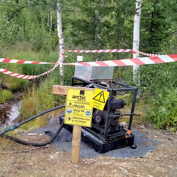 Maria Kristoffersson i grön fleecejacka och en pin dör det står ”uranbrytning nej tack”. Till vänster syns en avspärrad provborrning i skogen med skyltar och  varningar.