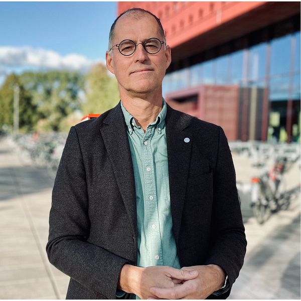 En person står framför en byggnad med röd fasad och cyklar parkering på bakgrunden. Till höger visas ett dokument med markerade rader som visar antalet sökande och antagna studenter för två olika terminer: HT2015 och HT2024. Texten anger ”sökande: 299” för HT2015 och ”sökande: 155” för HT2024.