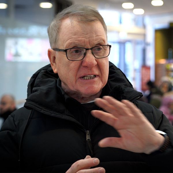 Man med grått hår och glasögon gör gest med händerna. Man brottar ner någon på golvet.