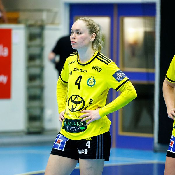Två handbollsspelare står på planen, klädda i gula matchtröjor med svarta detaljer. Den ena spelaren har nummer 4 på bröstet och den andra har nummer 20. De ser fokuserade ut och väntar på spelets fortsättning. Bakgrunden visar en sportarena med blått golv och publikområde.