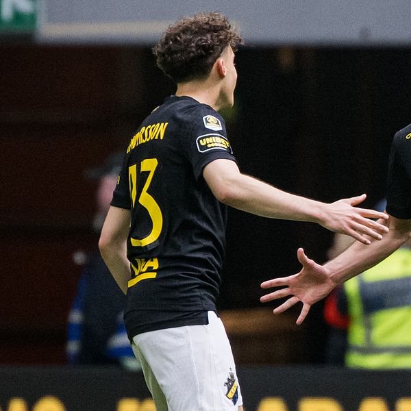 Två AIK-spelare på fotbollsplan med sträckta händer, publik och vakter i bakgrunden.
