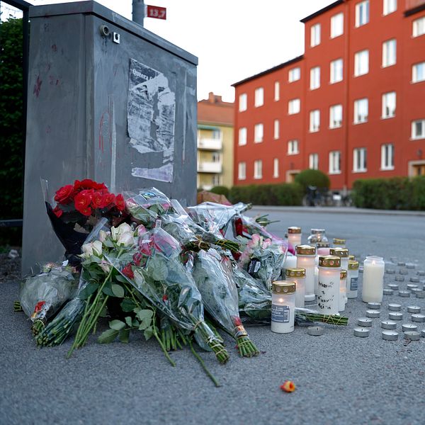 Blommor och tända ljus på trottoar, tre personer samlade i bakgrunden vid rött bostadshus.