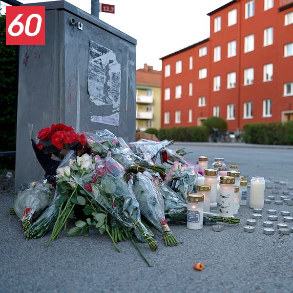 Blommor och tända ljus på trottoar, tre personer samlade i bakgrunden vid rött bostadshus.