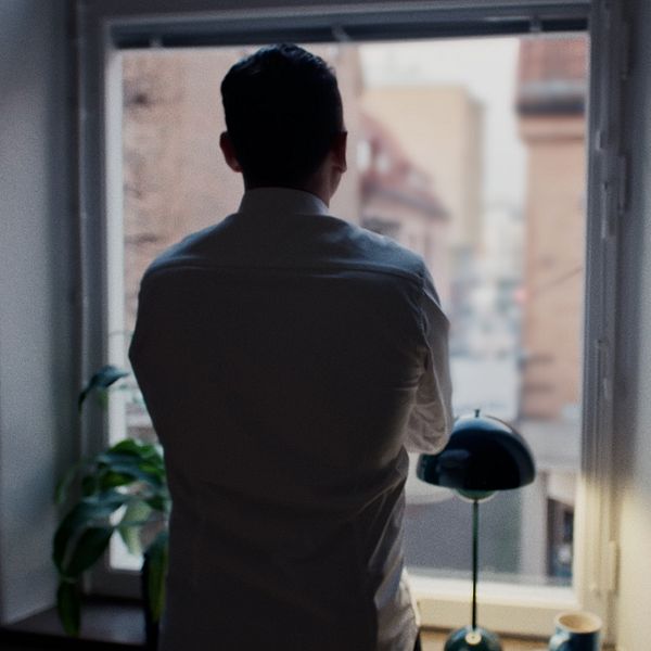 En delad bild: Till vänster syns en man bakifrån, stående vid ett fönster och tittar ut över en urban miljö. Till höger hålls en närbild av en person i en ljus kavaj, med en inomhusinredning i bakgrunden.