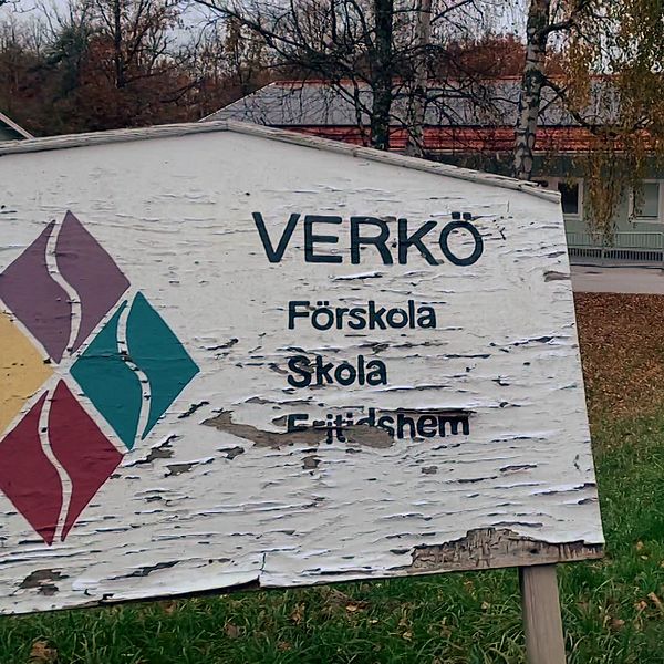 Skylt vid Verkø Förskola, Skola och Fritidhem med en suddig bakgrund av byggnaden och omgivande träd i höst. Skyltens text visar namnet på institutionen och dess verksamheter.