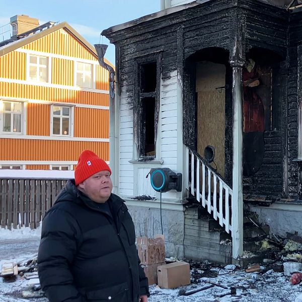 Man i röd mössa står vid brandhärjat hus med sotiga fasaden och bråte på marken.