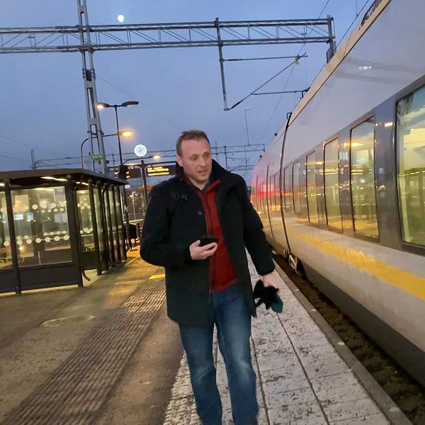 En person går längs en perrong bredvid ett Västtåg som står stilla på stationen. Tåget har gul och grå design och det är skymning utomhus. Plattformen har några lyktstolpar och en väntsal i bakgrunden.