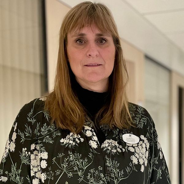 En person står i en korridor med glaspartier längs väggarna. Personen bär en blommig outfit och uttrycker en professionell attityd. Korridoren är belyst av mjukt ljus som kommer från fönstren, och i bakgrunden syns fler kontorsutrymmen.