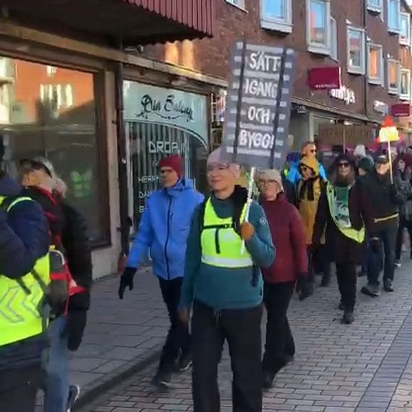 Människor deltar i en demonstration och går längs en gata, bärande på plakat. De flesta har på sig reflexvästar och olika kläder, och de syns representera en grupp med gemensamt syfte. I bakgrunden syns affärslokaler och stadsarkitektur.