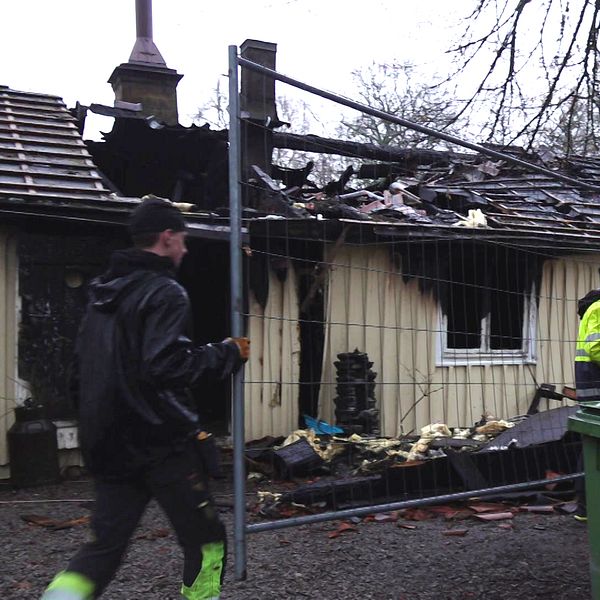 Räddningstjänst och räddningsarbetare inspekterar en brandhärjad byggnad med delvis förstört tak. En man i svart jacka går förbi med ett stängsel i bakgrunden, medan en annan man i räddningskläder arbetar i närheten av en grön soptunna. Rester av byggmaterial ligger utspridda på marken.