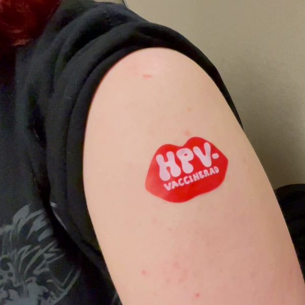 Överarm med klistermärke efter HPV-vaccination. Del av väntrum syns i bakgrunden.
