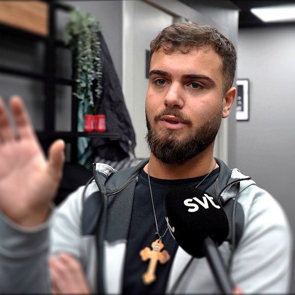 Person intervjuas av SVT inomhus, kors runt halsen, ytterligare person sitter i bakgrunden.